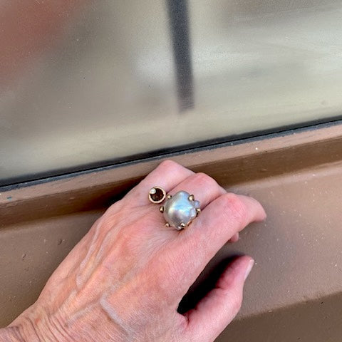 Bague ouverte et sculpturale en bronze. Ornée d'une grande perle baroque grise d'un côté et d'un diamant de l'autre. Bijou porté.