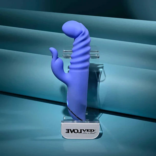 A purple curved silicone adult novelty device displayed upright on a clear stand with a white base labeled EVOLVED, set against a blue studio backdrop.