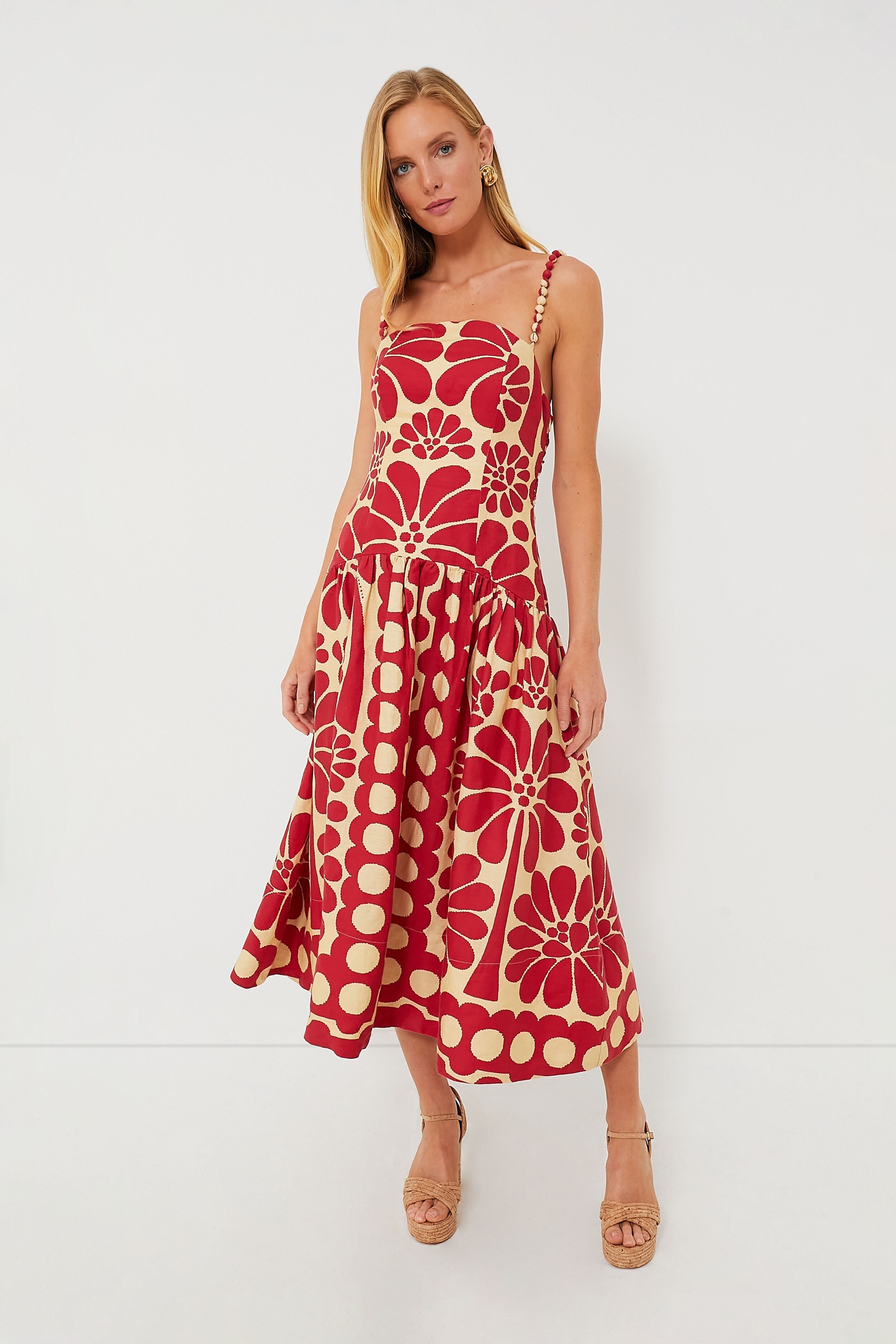 A woman wears a Farm Rio dress—a vibrant, sleeveless, knee-length style featuring red and beige floral and polka dot patterns—showcasing the brand's sustainable fashion.