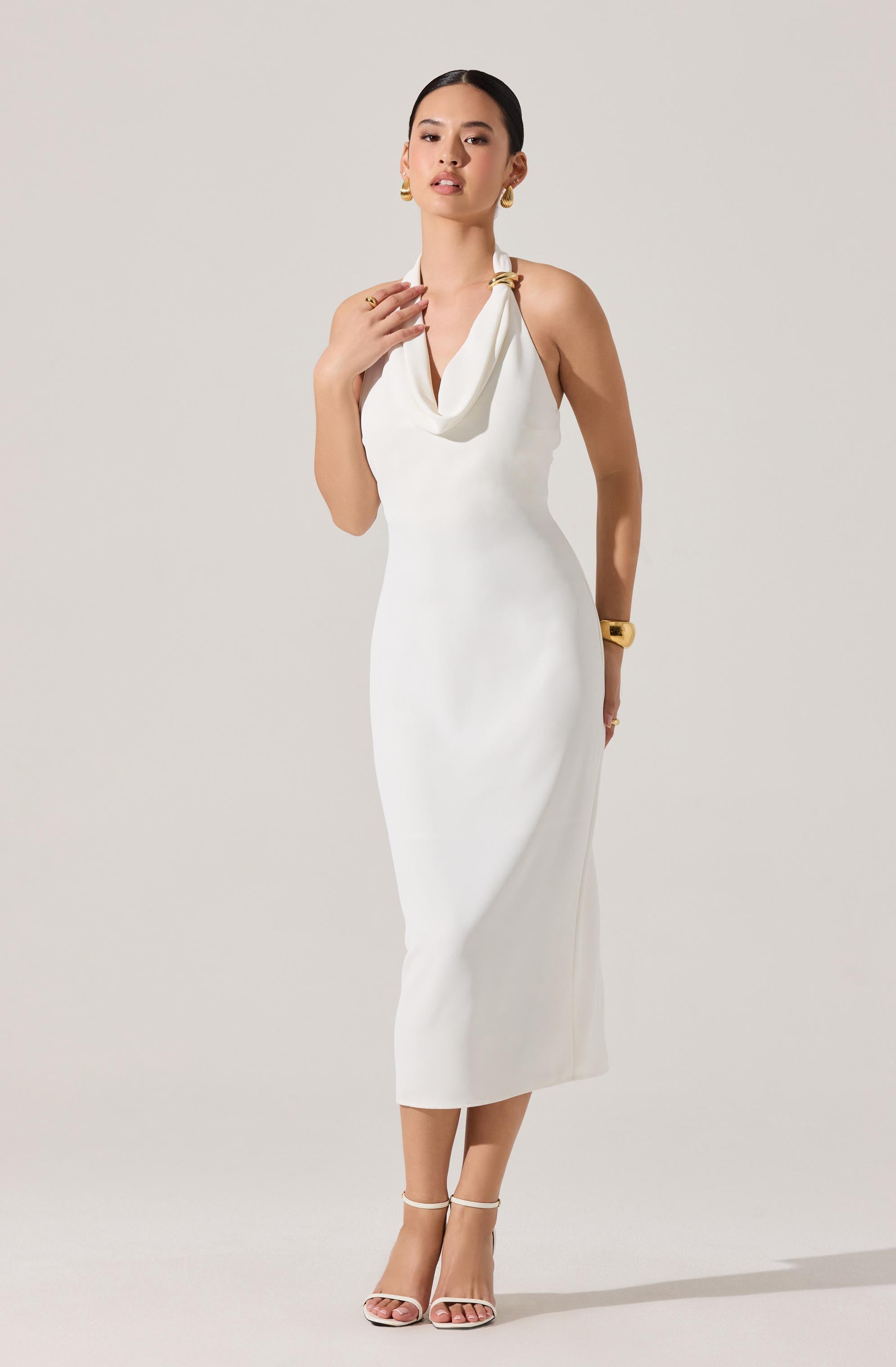A woman is wearing a sleeveless, fitted, white midi dress with a draped neckline. The dress has a sleek, elegant silhouette extending to her mid-calf. She accessorizes with gold hoop earrings, a gold bracelet on her right wrist, and a gold ring on her left hand. She completes her look with white strappy high-heeled sandals.